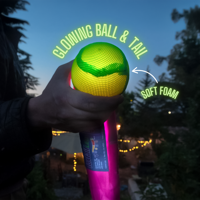 A hand holds a Foxtail LED outdoor game toy with a glowing yellow-green ball and tail labeled "GLOWING BALL & TAIL" and "SOFT FOAM." Trees and string lights at dusk set the scene for light-up nighttime play.