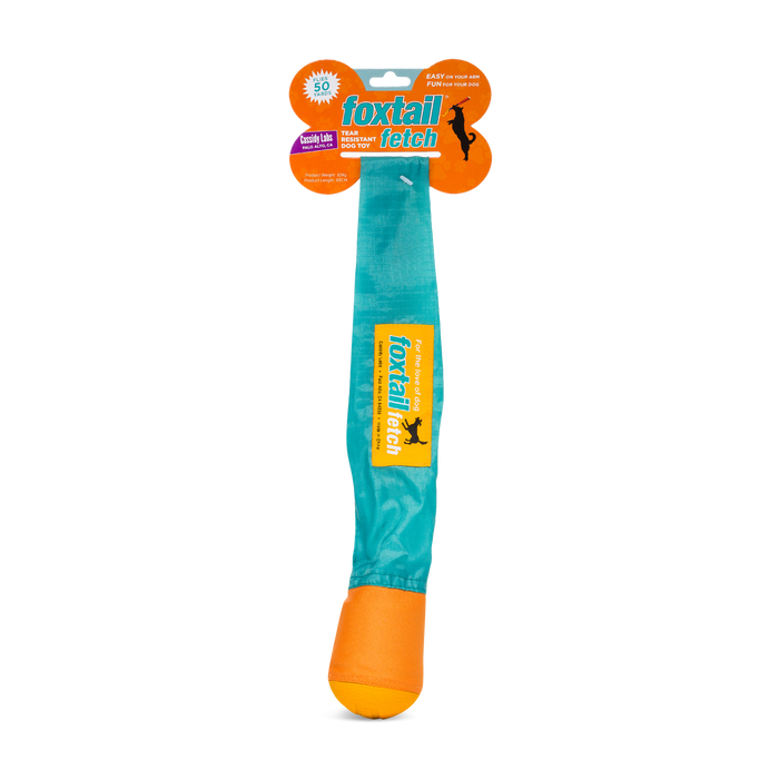 A Foxtail Fetch (Dog Toy) in its packaging, featuring a long teal tail attached to an orange rubber ball, designed for durable, slobber-free fetching and throwing games with dogs.